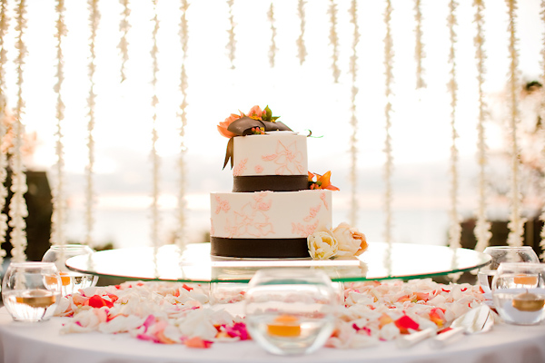 White two tiered round wedding cake with dark brown borders and orange floral designs as borders with orange, yellow, and ivory floral accents - Wedding cake surrounded by orange floating candles and rose petals - wedding photo by Michael Norwood Photography